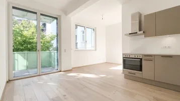 Heller offener Wohnbereich mit moderner Küchenzeile, Holzboden und Zugang zu einem Balkon.