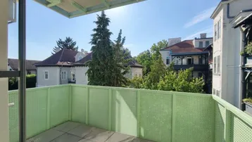 Sonniger Balkon mit grüner Brüstung, Fliesenboden und Blick auf die umliegenden Gebäude und Bäume.
