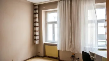 Helles Zimmer mit beigen Wänden, Holzfußboden und einem Fenster mit Vorhängen und Heizkörper.
