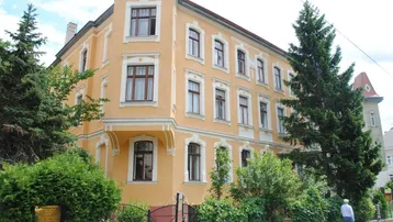 Historisches Gebäude mit gelber Fassade, vielen Fenstern und umgebender Bepflanzung.