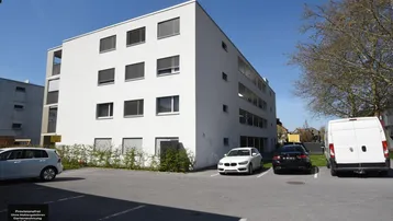 Modernes Mehrfamilienhaus mit weißer Fassade und Außenparkplätzen unter blauem Himmel.