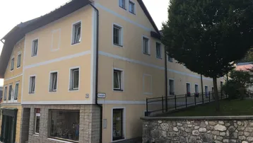 Gut erhaltenes Gebäude mit gelber Fassade, Satteldach und einer Rampe mit Handlauf zum erhöhten Grünbereich.