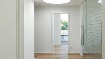 Heller Flur mit Holzboden, runden Deckenleuchten und einer Glaswand, die Tageslicht hereinlässt.
