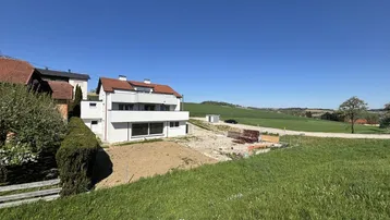 Weitläufige Ansicht des modernen Wohnhauses mit weißer Fassade und ländlicher Umgebung unter blauem Himmel.