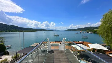 Panoramablick auf den See mit Segelbooten und Bergen, von einer modernen Holzterrasse mit Glasgeländer.