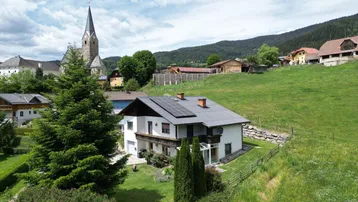 Luftaufnahme eines Hauses mit Solaranlage auf dem Dach und einer Kirche im Hintergrund, umgeben von grünen Hügeln.