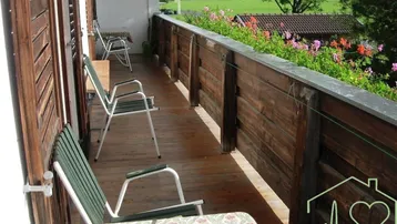 Langer Balkon mit Holzgeländer, Sitzgelegenheiten und Blick auf grüne Wiesen und Berge.