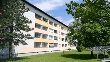 Mehrfamilienhaus mit grüner Rasenfläche und Bäumen im Vordergrund unter blauem Himmel.