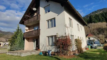 Mehrstöckiges Wohnhaus mit Balkonen und Gartenbereich in ländlicher Umgebung und Bergblick.