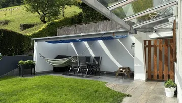 Überdachte Terrasse mit Gartenmöbeln und Hängematte, umgeben von Grünflächen und Hügeln.