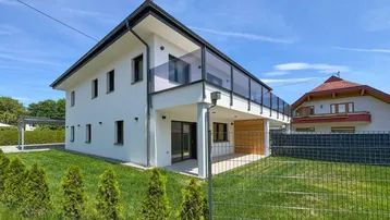 Modernes Wohngebäude mit weißer Fassade, großem Garten und Balkon mit Glasgeländer.