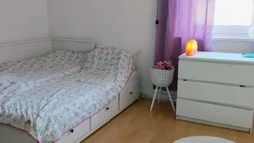 Helles Schlafzimmer mit Holzboden, weißer Kommode und lila Vorhängen am Fenster.