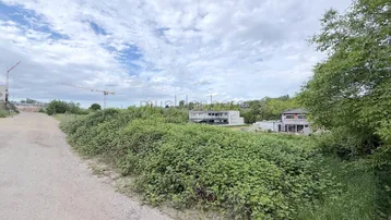 Blick auf ein unbebautes Grundstück mit umliegender Vegetation und fernen Gebäuden unter bewölktem Himmel.