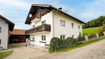 Weißes Landhaus mit Holzbalkonen und gepflasterter Auffahrt, umgeben von grüner Landschaft.