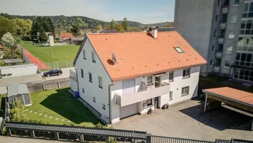 Einfamilienhaus mit rotem Ziegeldach, Carport und angrenzendem Sportplatz in ländlicher Umgebung.