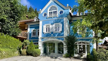 Historische Villa mit auffälliger blauer Fassade und weißen Fensterläden, umgeben von Grün.