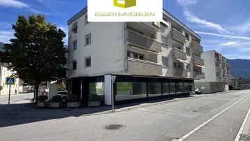 Mehrstöckiges Gebäude mit Geschäftsflächen im Erdgeschoss und Wohnungen darüber, an einer Straße gelegen.