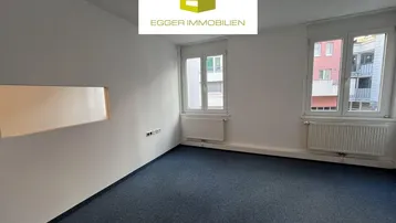 Helles Zimmer mit blauem Teppichboden und zwei Fenstern, ideal als Büro oder Schlafzimmer.