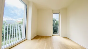 Helles, leeres Zimmer mit großen Fenstern, Parkettboden und Zugang zu einem französischen Balkon.
