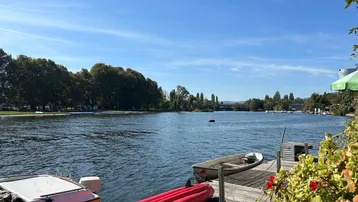 Seeufer mit Booten und Steg unter blauem Himmel, umgeben von grünen Bäumen.