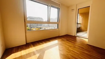 Helles Zimmer mit Parkettboden und großem Fenster, das viel Tageslicht hereinlässt und einen Stadtblick bietet.
