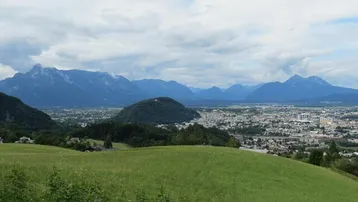 Weitläufiger Blick über eine grüne Landschaft mit einer Stadt im Tal und majestätischen Bergen im Hintergrund.