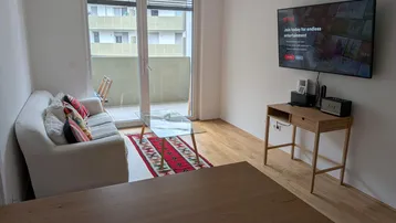 Helles Wohnzimmer mit Holzboden, Sofa, Fernseher und Zugang zu einem Balkon.