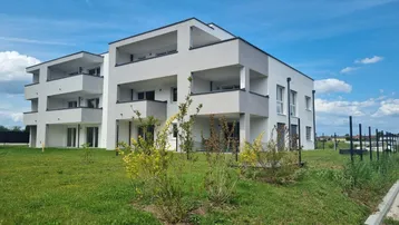 Modernes Mehrfamilienhaus mit weißer Fassade und Balkonen, umgeben von Grünflächen unter blauem Himmel.