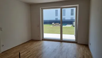Helles, leeres Zimmer mit Holzboden und großem Fenster mit Zugang zum Garten.