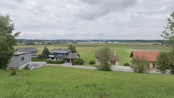 Blick auf eine ländliche Umgebung mit Wohnhäusern, weiten Feldern und einem Dorf in der Ferne unter bewölktem Himmel.