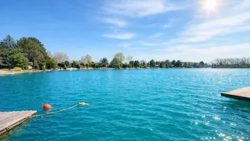 Weitläufiger Blick über den See mit einem Holzsteg im Vordergrund an einem sonnigen Tag.