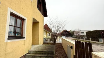 Seitenansicht des Hauses mit gepflastertem Weg und Holzzaun unter bewölktem Himmel.