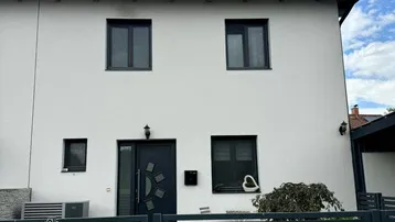 Moderne weiße Hausfassade mit dunklen Fenstern und Eingangstür, umgeben von einem Zaun.