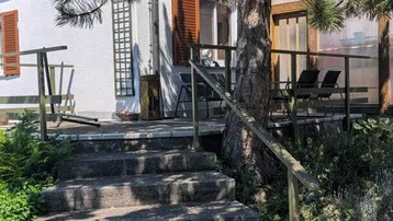 Eine Außentreppe führt zu einer erhöhten Holzterrasse mit Sitzgelegenheiten und einem großen Baum.