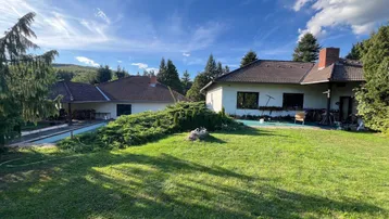 Weitläufiger Gartenbereich mit zwei Wohnhäusern und einem Pool im Hintergrund unter blauem Himmel.