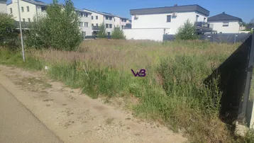 Unbebautes Grundstück mit hohem Gras und umliegenden Wohngebäuden unter blauem Himmel.