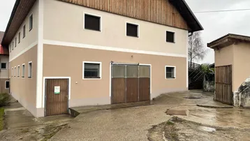 Gebäudeaußenansicht mit beiger Fassade, Holzverkleidung im oberen Bereich und großen Holztoren, umgeben von einem gepflasterten Hof.
