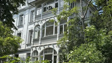 Historisches Gebäude mit eleganter Fassade und großen Fenstern, umgeben von üppigem Grün und Bäumen.