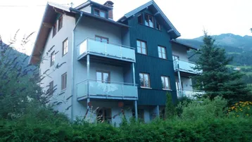 Modernes Mehrfamilienhaus mit Balkonen und Bergblick, umgeben von grünen Hecken und Bäumen.