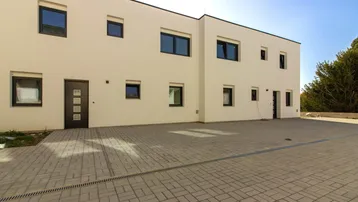 Modernes Mehrfamilienhaus mit heller Fassade und gepflastertem Vorplatz unter blauem Himmel.
