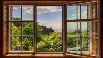 Fenster mit Blick auf grüne Bäume und eine weite Landschaft unter blauem Himmel.