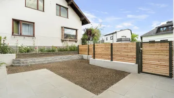Gepflegter Außenbereich mit gepflasterter Fläche, Kiesbeet und modernem Holzzaun vor einem Wohnhaus.