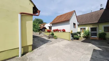 Innenhof zwischen zwei Gebäuden mit gepflastertem Bereich und Blumenbeeten unter blauem Himmel.
