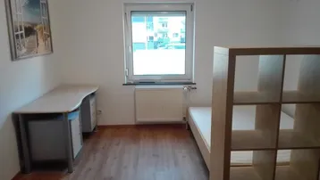 Helles Zimmer mit Fenster, Schreibtisch, Bettrahmen und Regal, ideal als Schlaf- oder Arbeitsbereich.