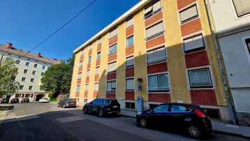 Mehrstöckiges Wohngebäude mit gelb-roter Fassade und parkenden Autos auf der Straße unter blauem Himmel.