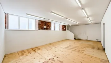 Großer, leerer Raum mit Sperrholzfußboden, weißen Wänden und einer Akzentwand aus Ziegeln, beleuchtet von Leuchtstoffröhren.