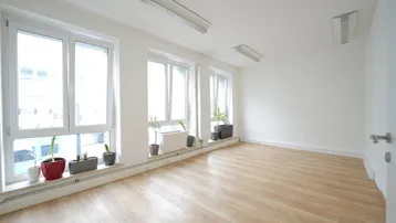 Heller Büroraum mit großen Fenstern, die viel Tageslicht hereinlassen, und hellem Laminatboden.