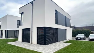Moderne Doppelhaushälfte mit weißer und grauer Fassade, umgeben von Rasenflächen und einer gepflasterten Terrasse.