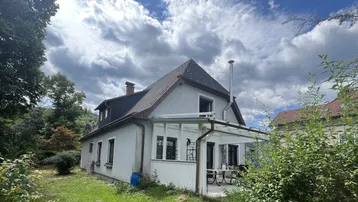 Hausfassade mit überdachter Terrasse und Gartenbereich unter bewölktem Himmel.