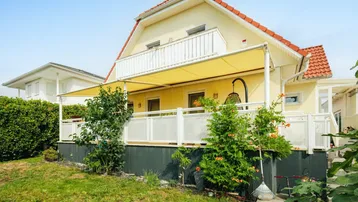 Gelbes Einfamilienhaus mit rotem Ziegeldach, Balkon und überdachter Terrasse im Grünen.